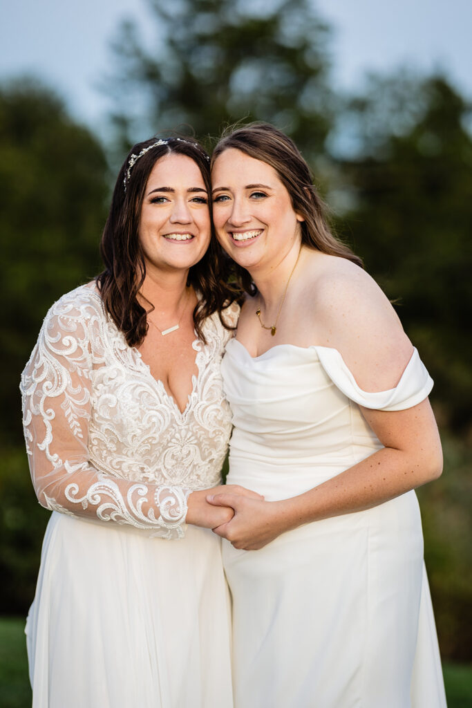 A close-up of two brides embracing and smiling at the camera, one in a lace-detailed dress and the other in an off-shoulder gown, sharing a joyful moment.