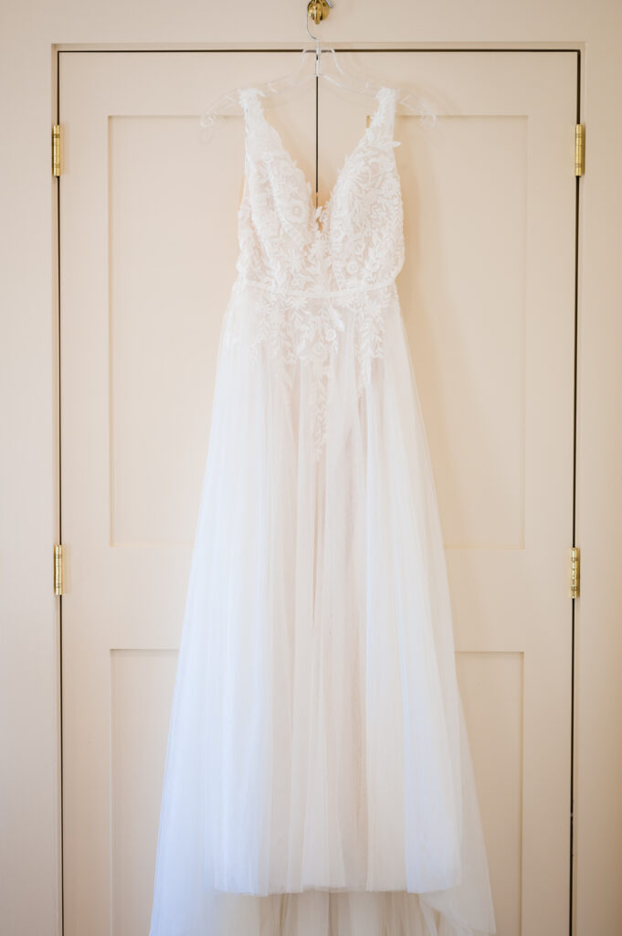A white lace and tulle wedding dress hanging on a clear hanger against a beige door.