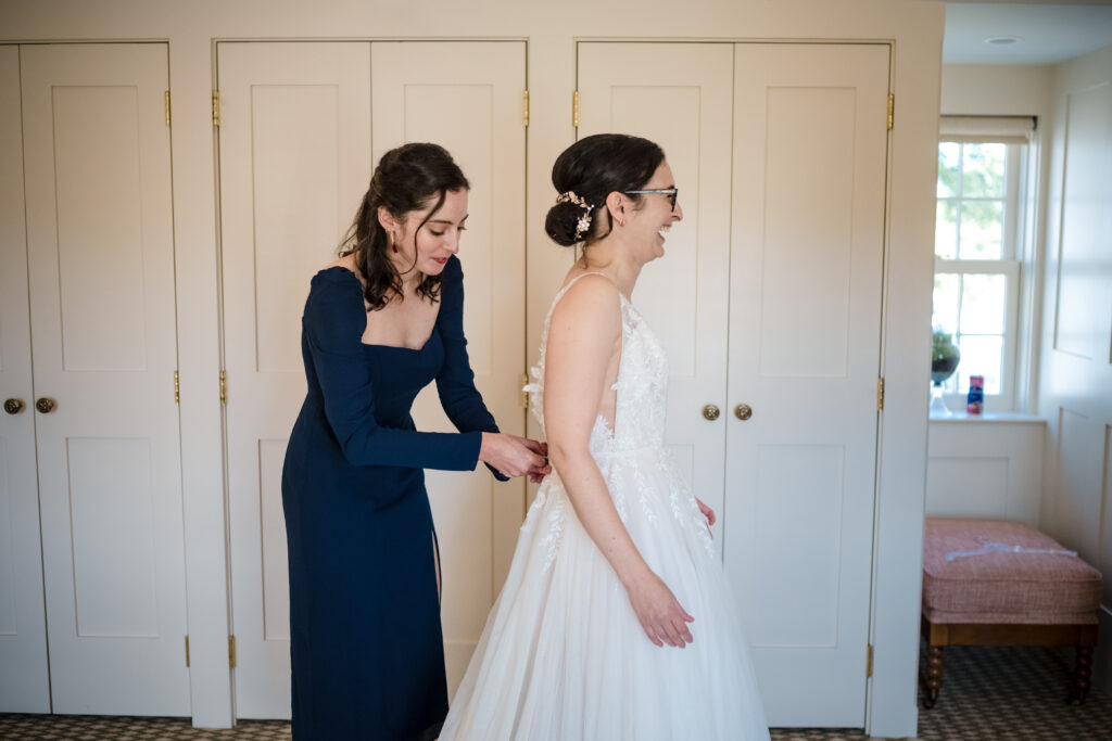 A woman in a dark dress helps another woman in a white wedding gown button the back of her dress in a well-lit room.