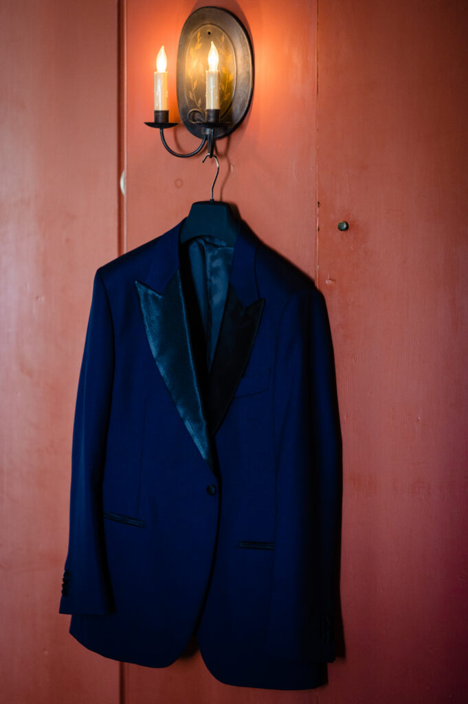 A dark blue tuxedo jacket with black satin lapels hangs on a black hanger beneath a lit wall sconce against a reddish-brown wall.