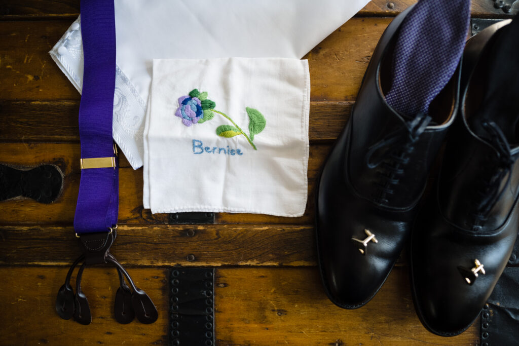 A pair of black dress shoes, a purple suspenders, and a white handkerchief with a flower and the name "Bernie" embroidered, all laid out on a wooden surface.