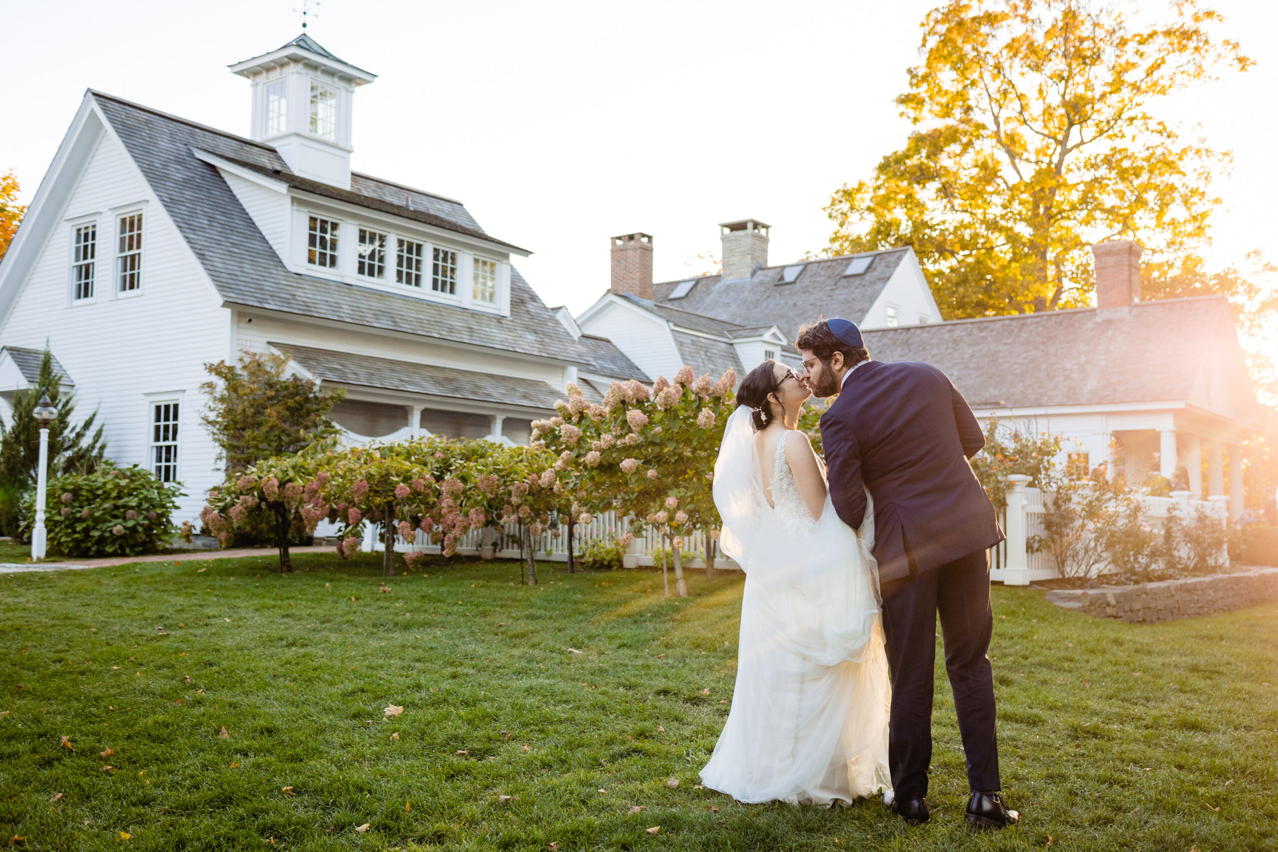 A bride and groom walk together on grass outside a large white house with blooming hydrangeas, trees, and sunlight in the background.
