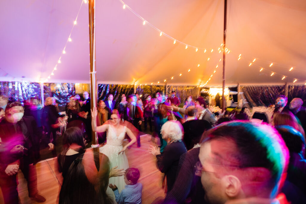 A group of people gather and dance under string lights in a tent at a wedding reception. The image is blurred, capturing motion and colorful lighting.