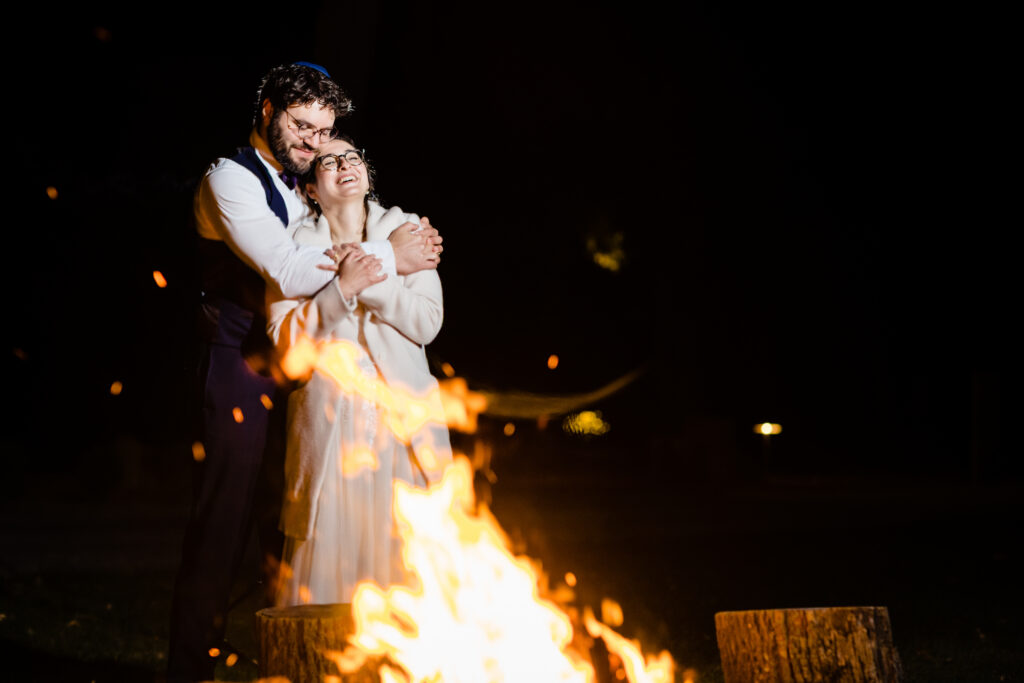 A couple dressed in formal attire stands outdoors at night, smiling and embracing near a large campfire with glowing flames.