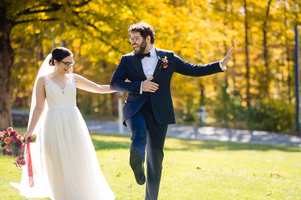 A bride and groom walk outside on bright green grass, smiling and holding hands. The groom kicks up one leg playfully. Autumn trees with yellow leaves are in the background.