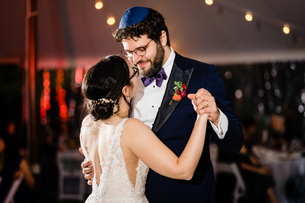 A bride and groom dance closely at their wedding reception under string lights. The groom wears a blue yarmulke and suit; the bride wears a white lace dress with an open back.